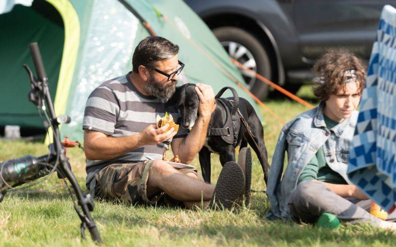 Man eating next to his dog, sitting outside a tent.