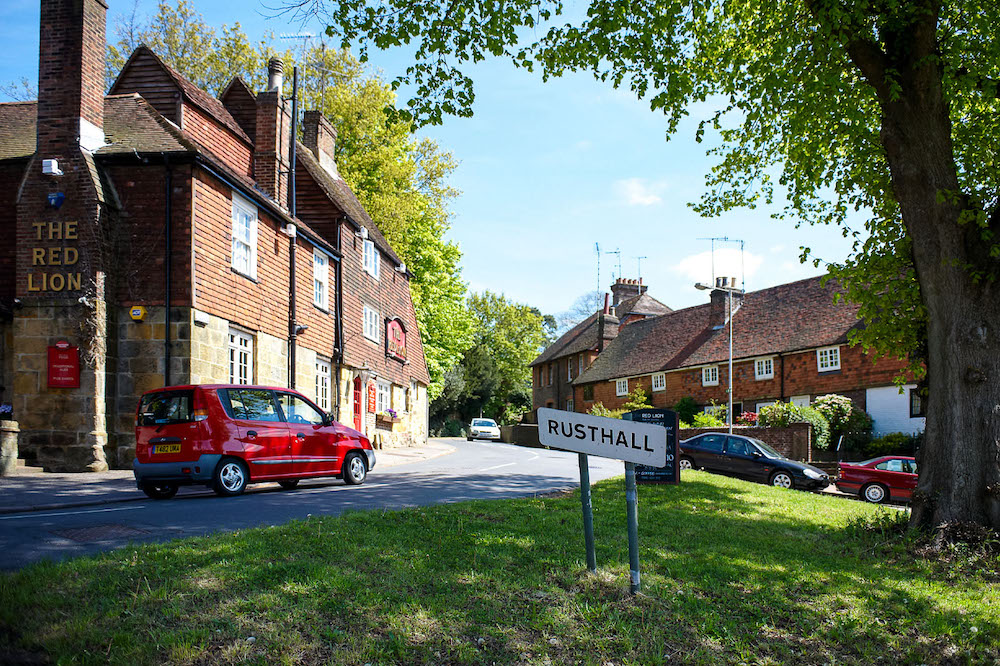 A street in Rusthall with greenery framing the image with the village sign of Rusthall in the centre, a pub to the left and houses to the right