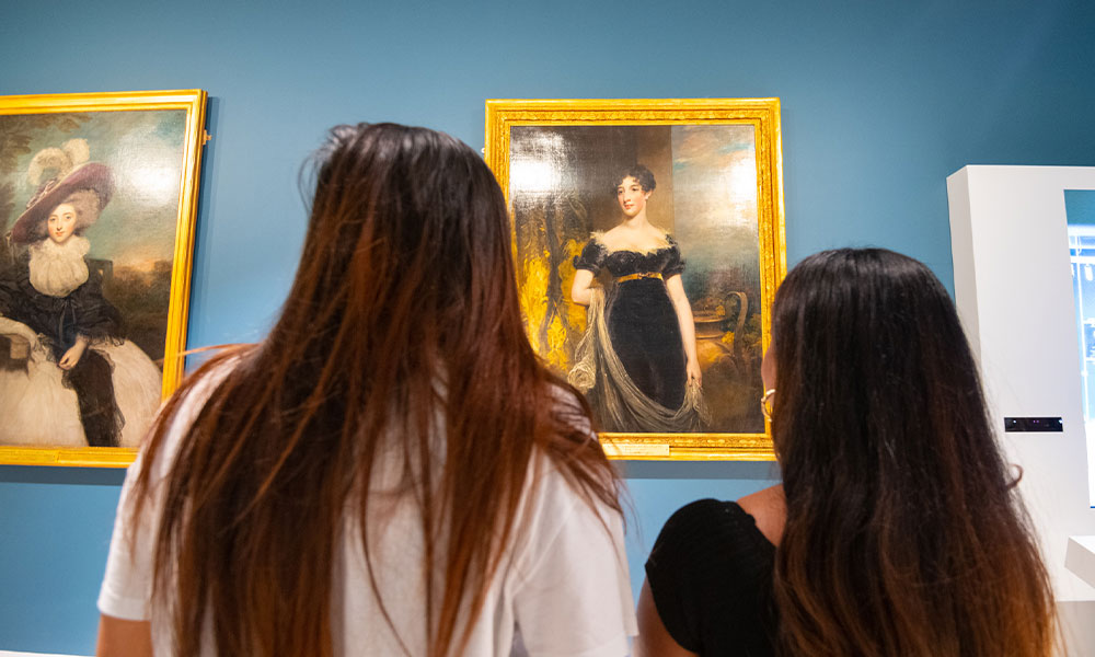 two ladies looking the portraits in the Camden Room at the Amelias Scott. The portrait are placed on a Royal Blue wall