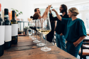 group of people tasting wine. The foreground includes a line of wine glasses and various bottles with labels covered.