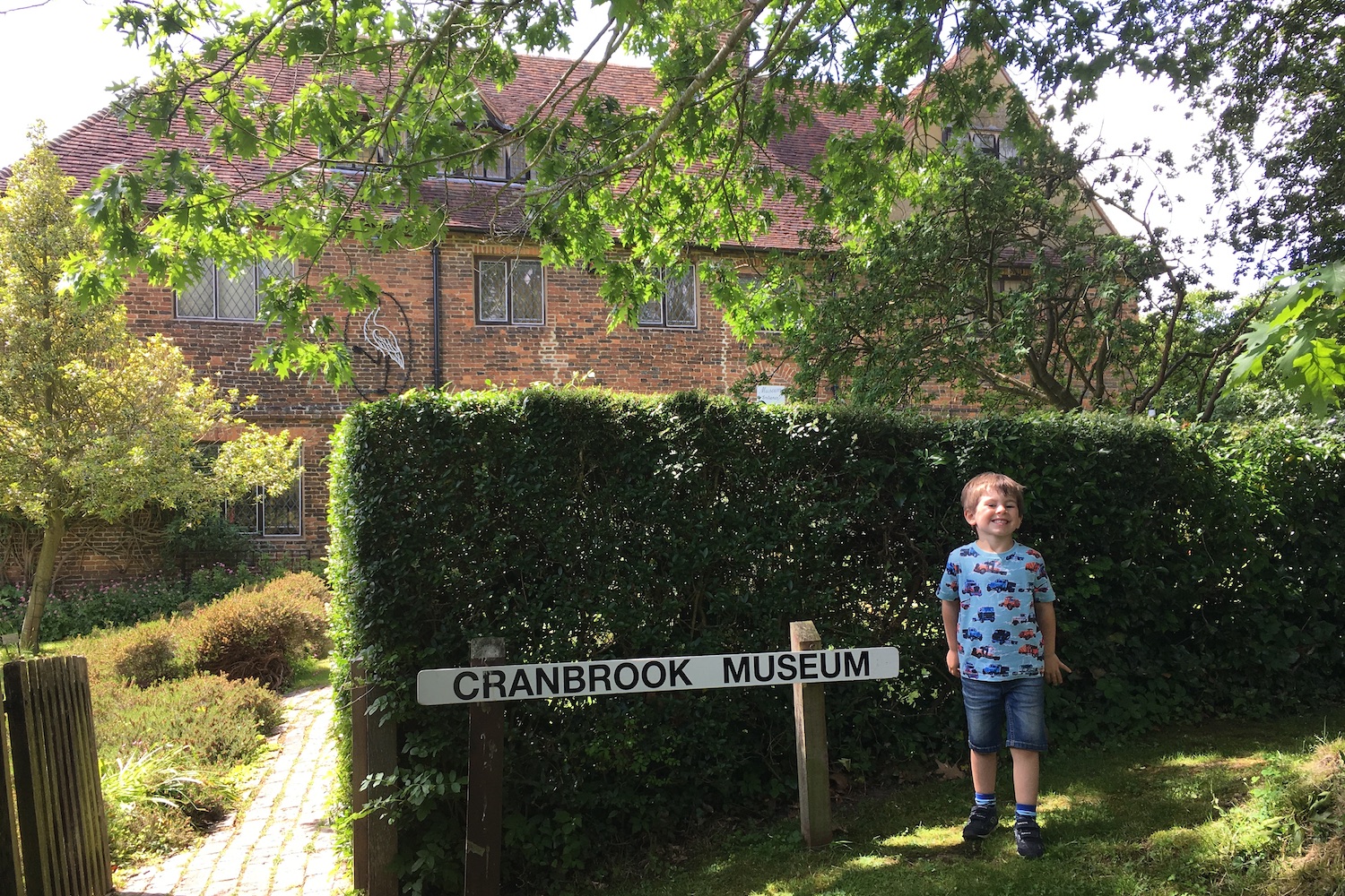 A young boy standing in front of a building smiling standing next to a sign that reads Cranbrook Museum
