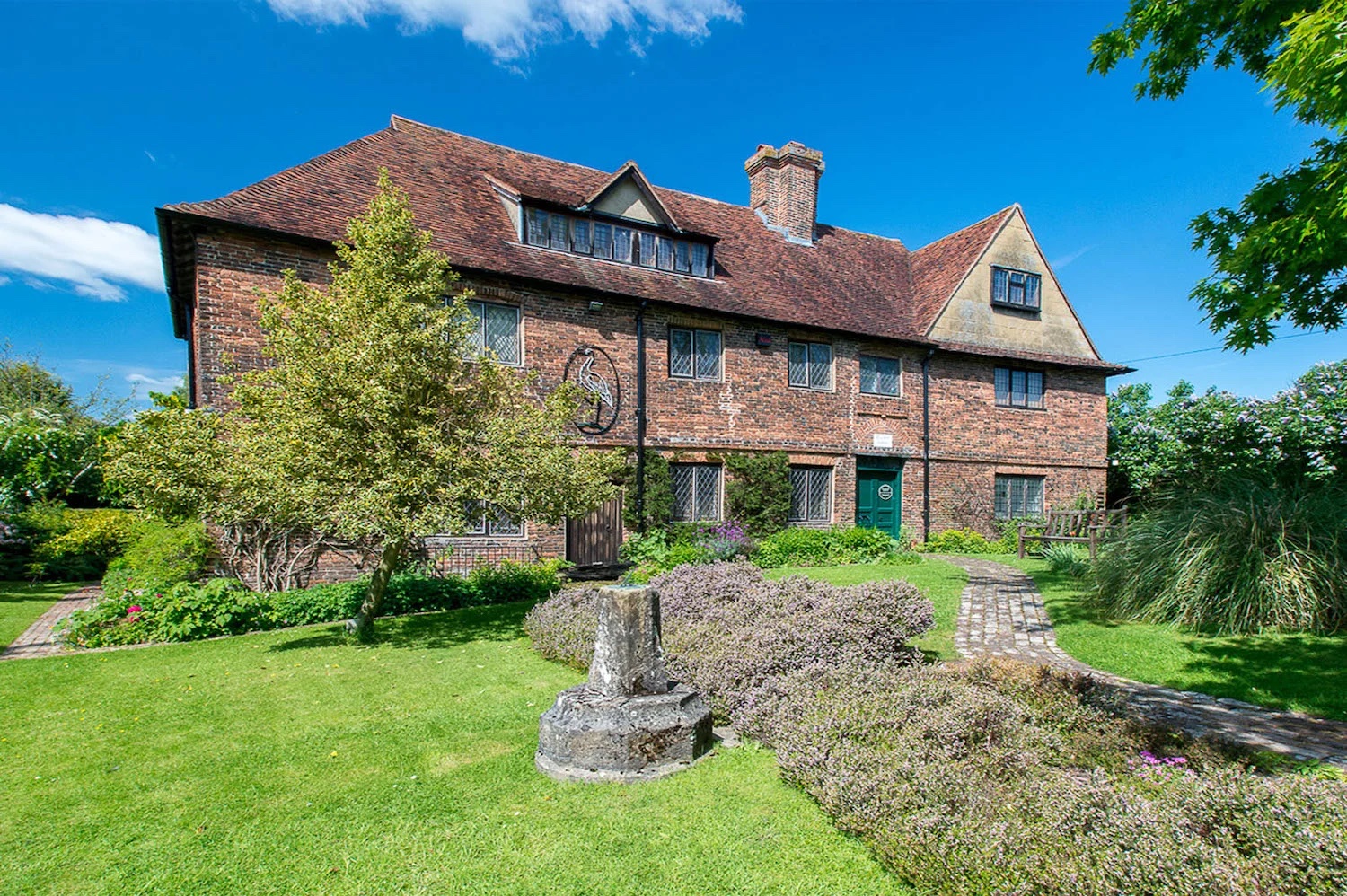 An old 15th century building which is now Cranbrook Museum with a winding pathway leading up to it with green grass, a tree and flowers decorated in front of it