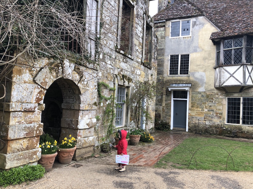 A young child dressed in a red coat holding a trail leaflet standing in front of Scotney Castle